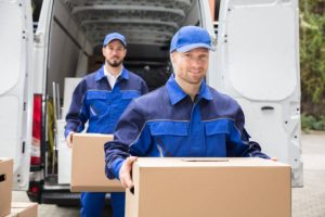 Close-up Of Two Young Delivery Men Carrying Cardboard Box In Front Of Truck